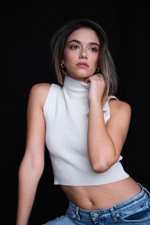 Fashion portrait of model in white turtleneck crop top and jeans against black background.