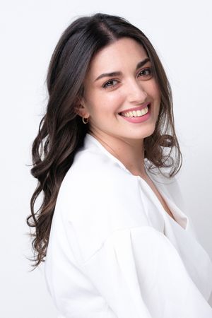 Professional headshot of a woman with wavy dark hair wearing a white blouse against white background.