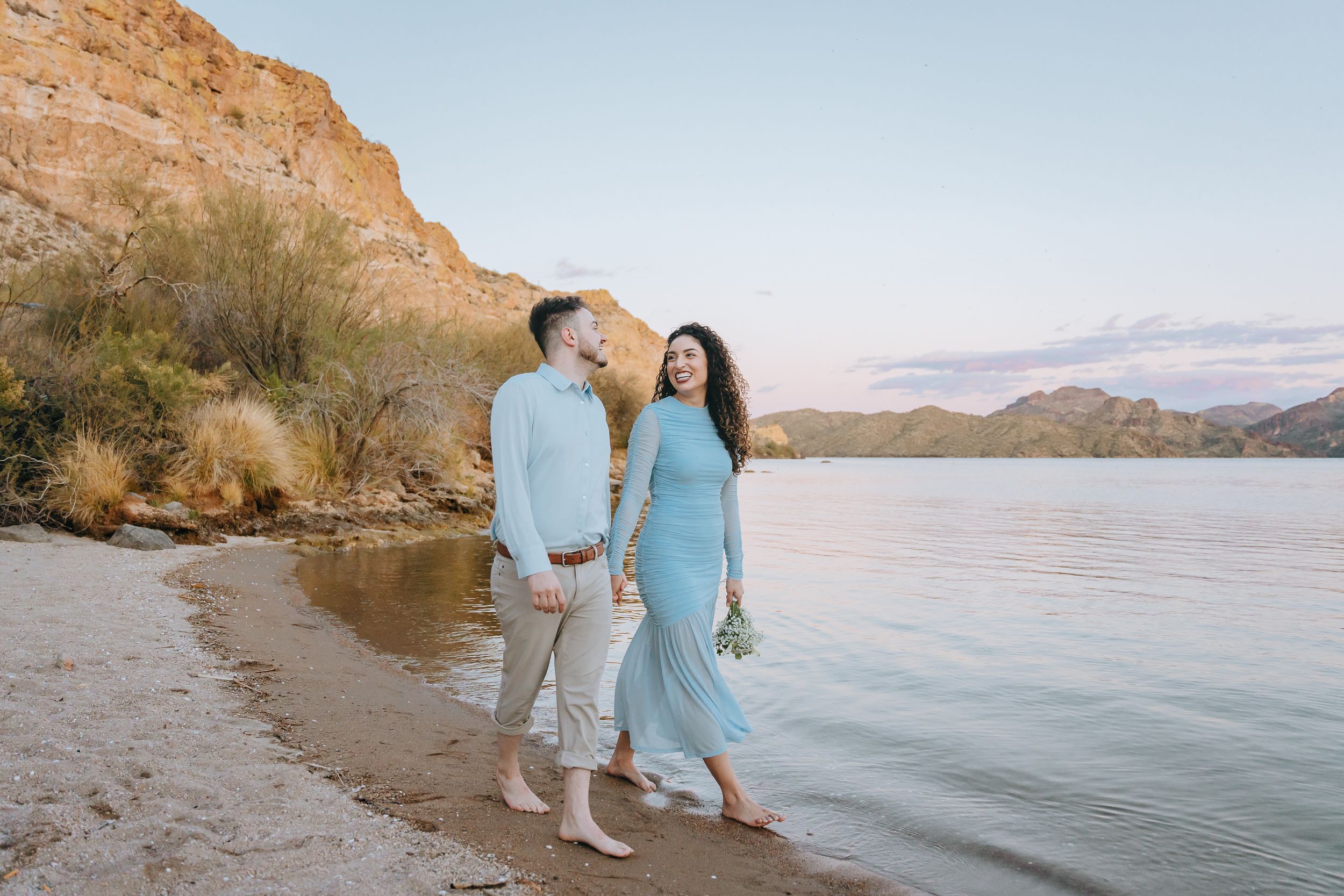 Couples walking along rocky shoreline with mountain and lake views at sunset.
