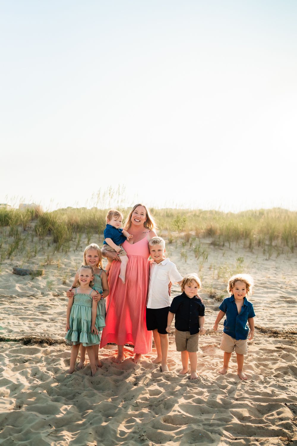 A group in coordinated outfits poses together on a sandy beach at sunset with grass dunes in the background.