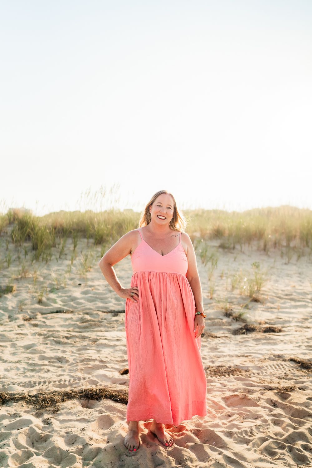 Person in coral pink dress photographed on sandy beach with dunes at golden hour.