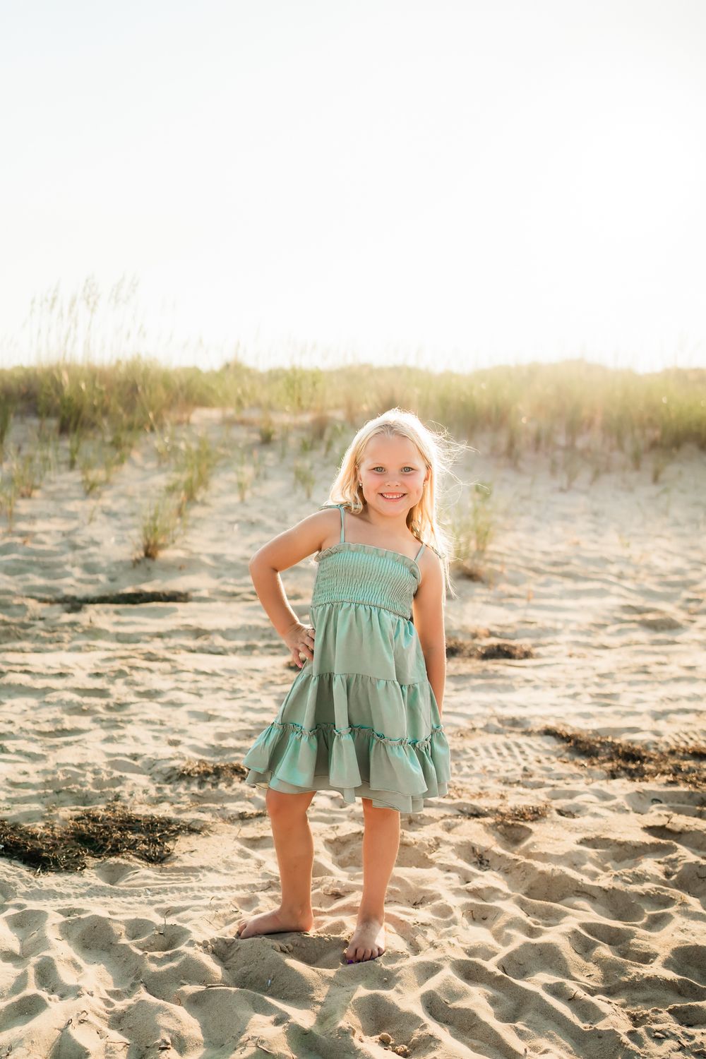 Young child in a mint green dress plays and poses on a sandy beach during golden hour.