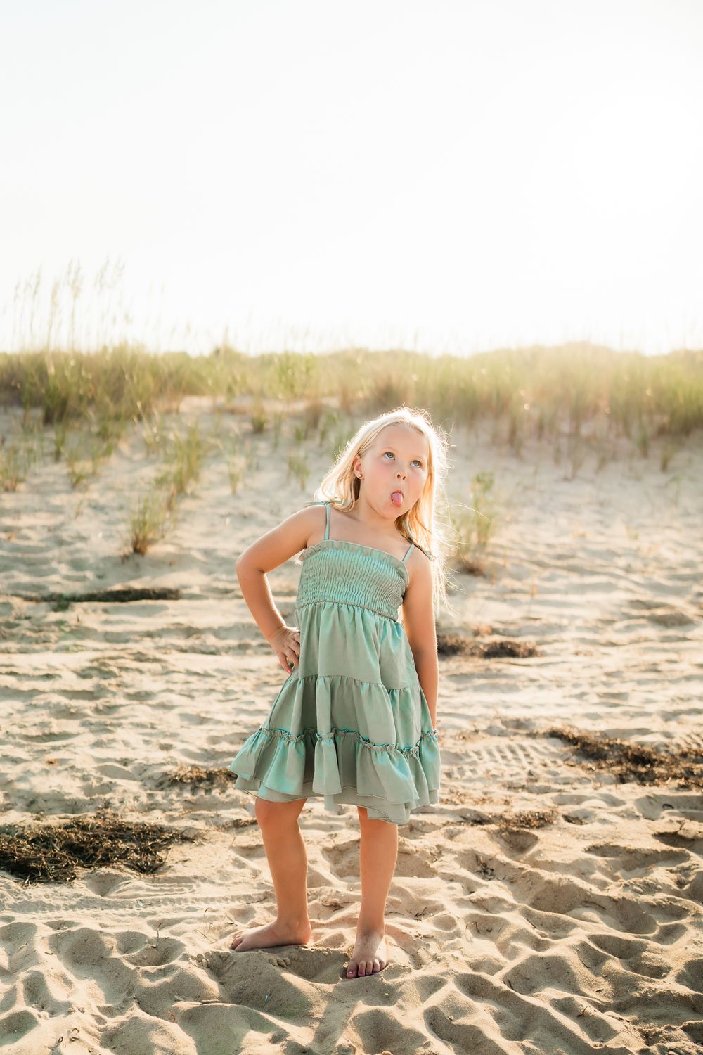 A small child wearing a mint green sundress stands playfully on a sandy beach at sunset.