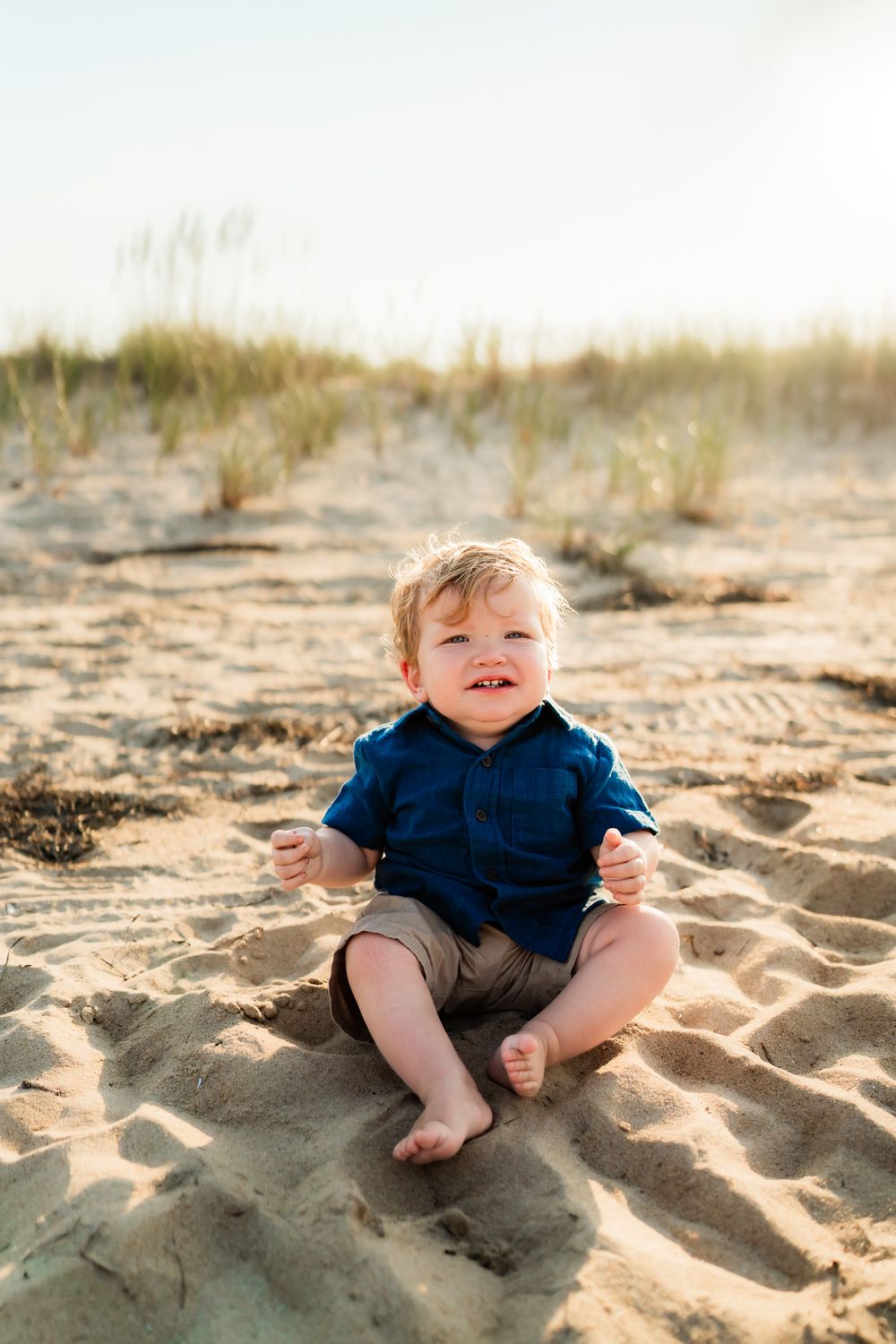 Small child wearing navy blue outfit sits happily in the sand at the beach.