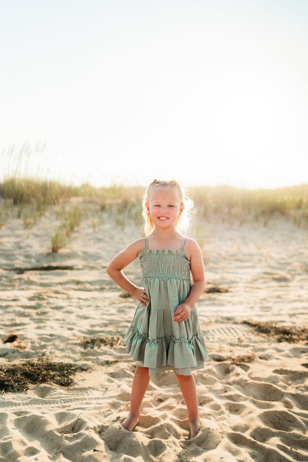 Sweet toddler in sage green ruffled romper plays joyfully on sandy beach at golden sunset.