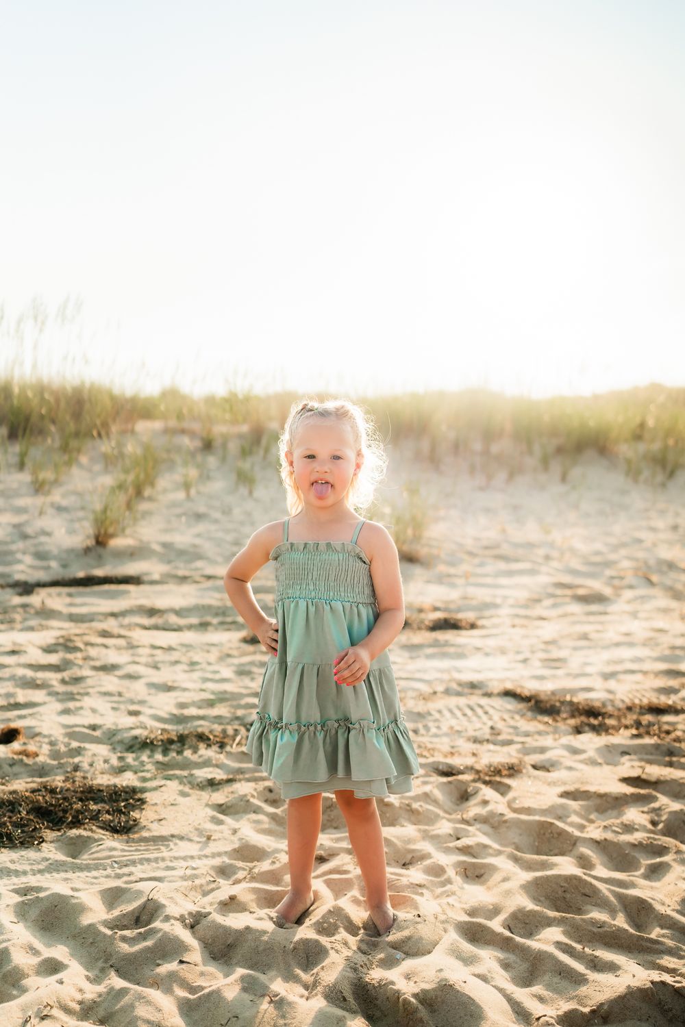A child in a mint green sundress stands confidently on a sandy beach at sunset.