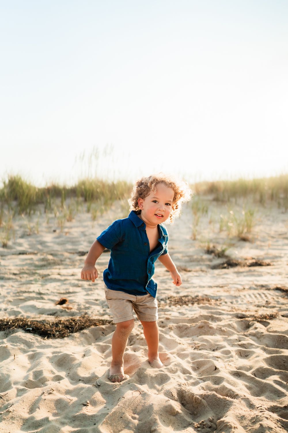 A small child plays joyfully on a sandy beach wearing a navy shirt and khaki shorts on a sunny afternoon.
