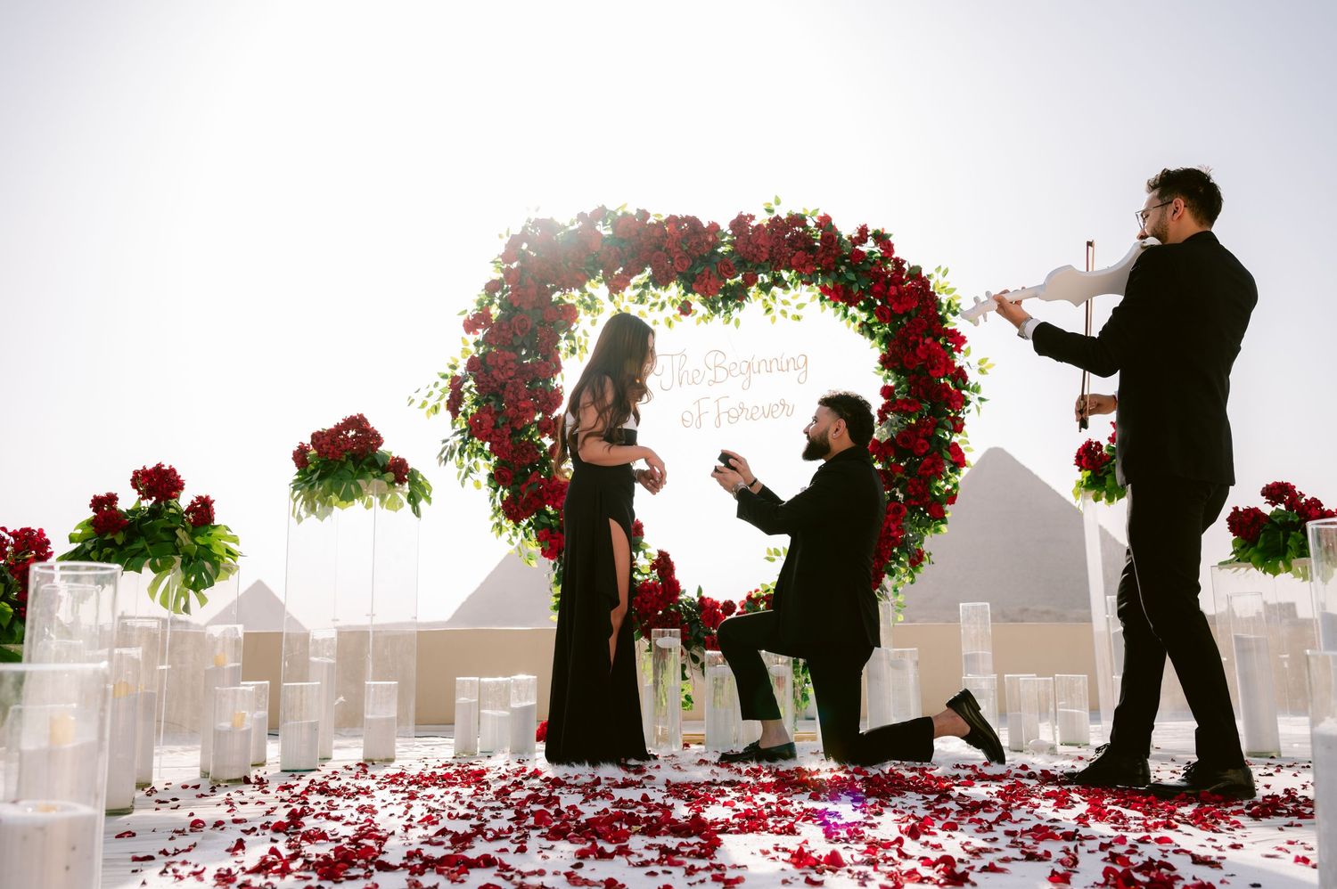 A romantic marriage proposal scene under a floral arch decorated with red and white flowers and scattered rose petals.