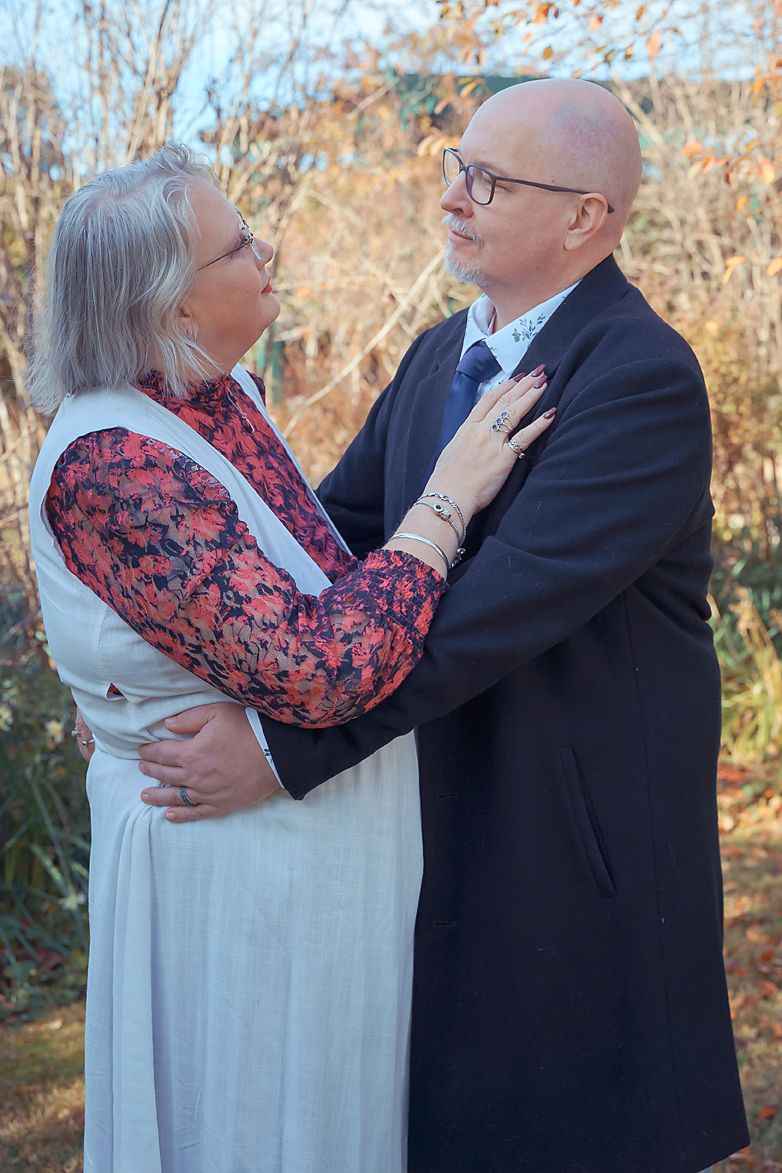 An elderly couple in formal attire share an intimate embrace outdoors during autumn with bare trees in the background.