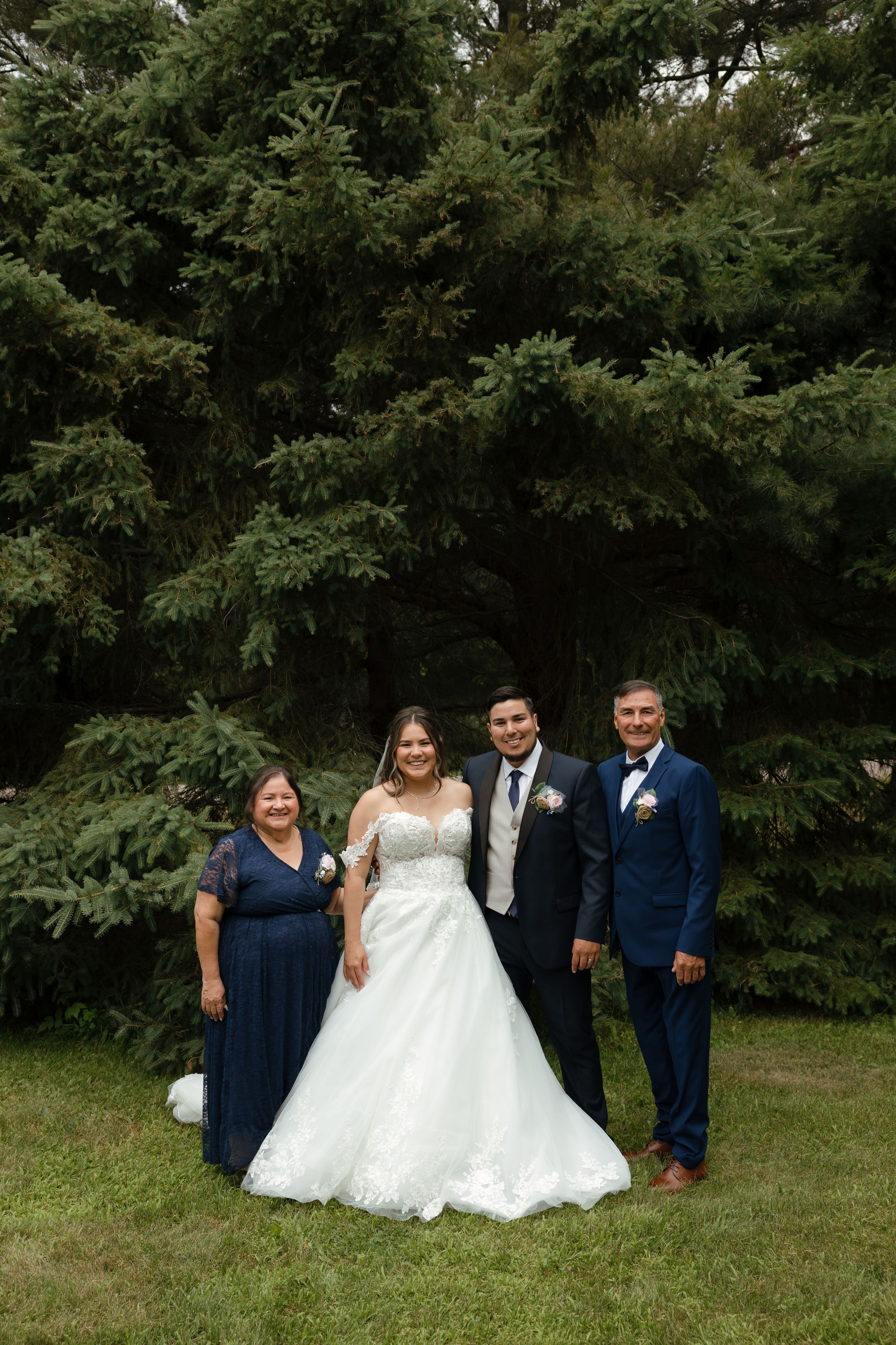 Wedding party poses on grass with bridesmaids in navy dresses and groomsmen in blue suits against evergreen backdrop.