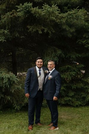 Two men in navy suits stand together on grass with trees in the background during a formal outdoor event.