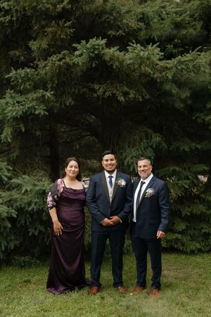 A wedding party in navy suits and purple dresses poses together in front of evergreen trees.