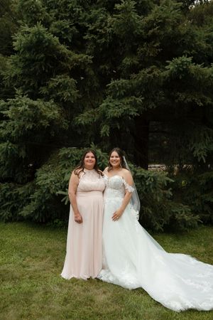 Two women wearing pink and white dresses pose together on a grassy lawn with trees in the background.