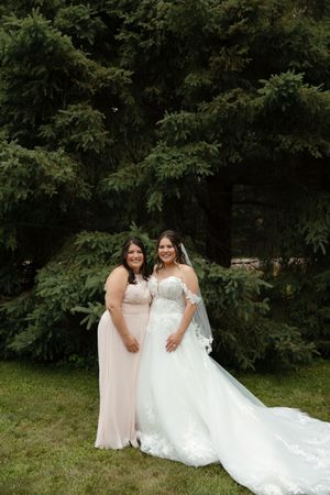 Two people in wedding attire sit together on grassy lawn with trees in background.