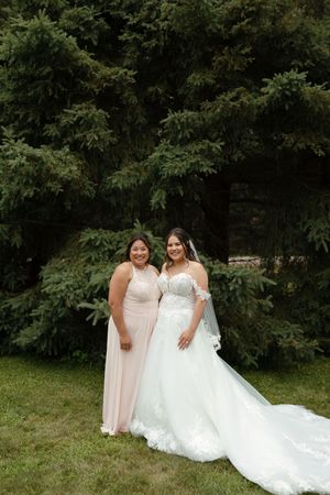 Two figures stand together outdoors in formal attire with long flowing white gown and blush pink dress against leafy backdrop.