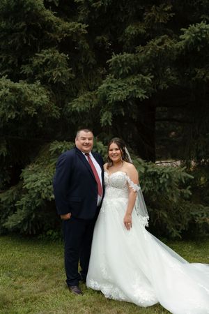 A couple in wedding attire poses together outdoors against a backdrop of trees beside a grassy area.