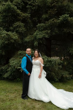 Two wedding figures in formal attire pose playfully on green grass with trees in the background.