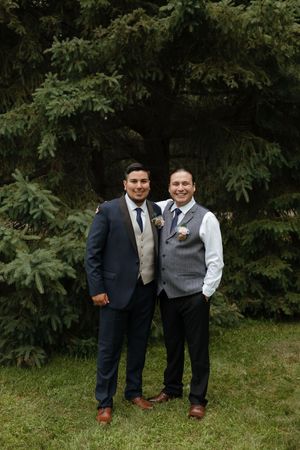 Two people wearing formal attire pose together for photos outdoors against a backdrop of green trees and foliage.