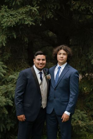 Two formally dressed friends in suits standing together against a dark green foliage background, sharing candid moments.
