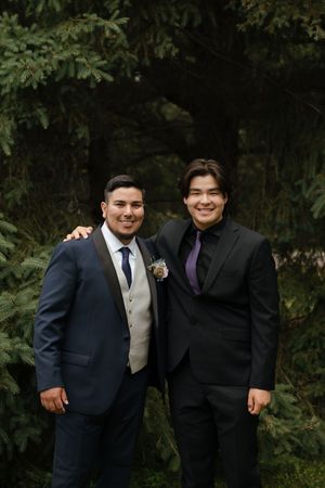 Two people in formal attire pose together outdoors against a backdrop of green foliage.
