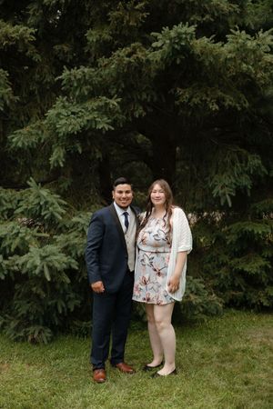 A couple stands together outdoors in formal attire against a background of lush green foliage and trees.