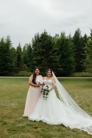 Two people in wedding attire pose together on a grassy field with evergreen trees in the background.
