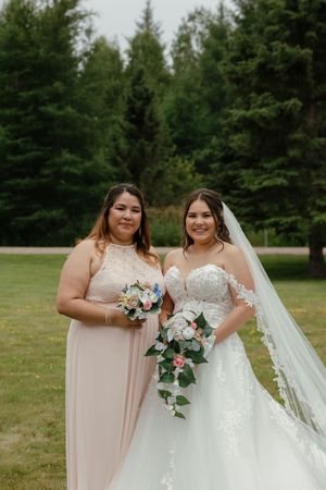 Two people in formal wedding attire pose outdoors with floral bouquets against a backdrop of evergreen trees.