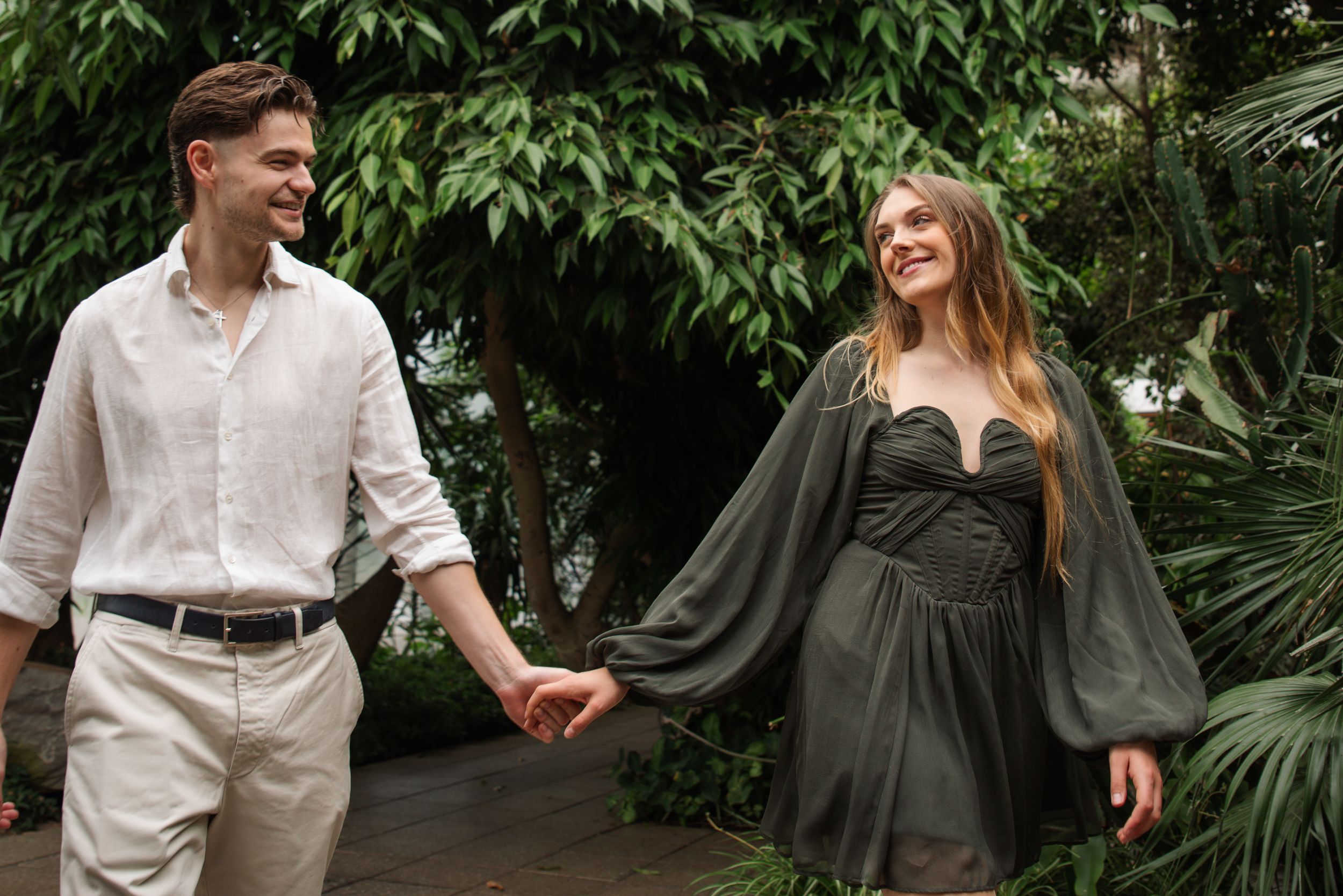 Happy couple holding hands and walking through a lush tropical garden, man in linen shirt and woman in elegant gray dress.