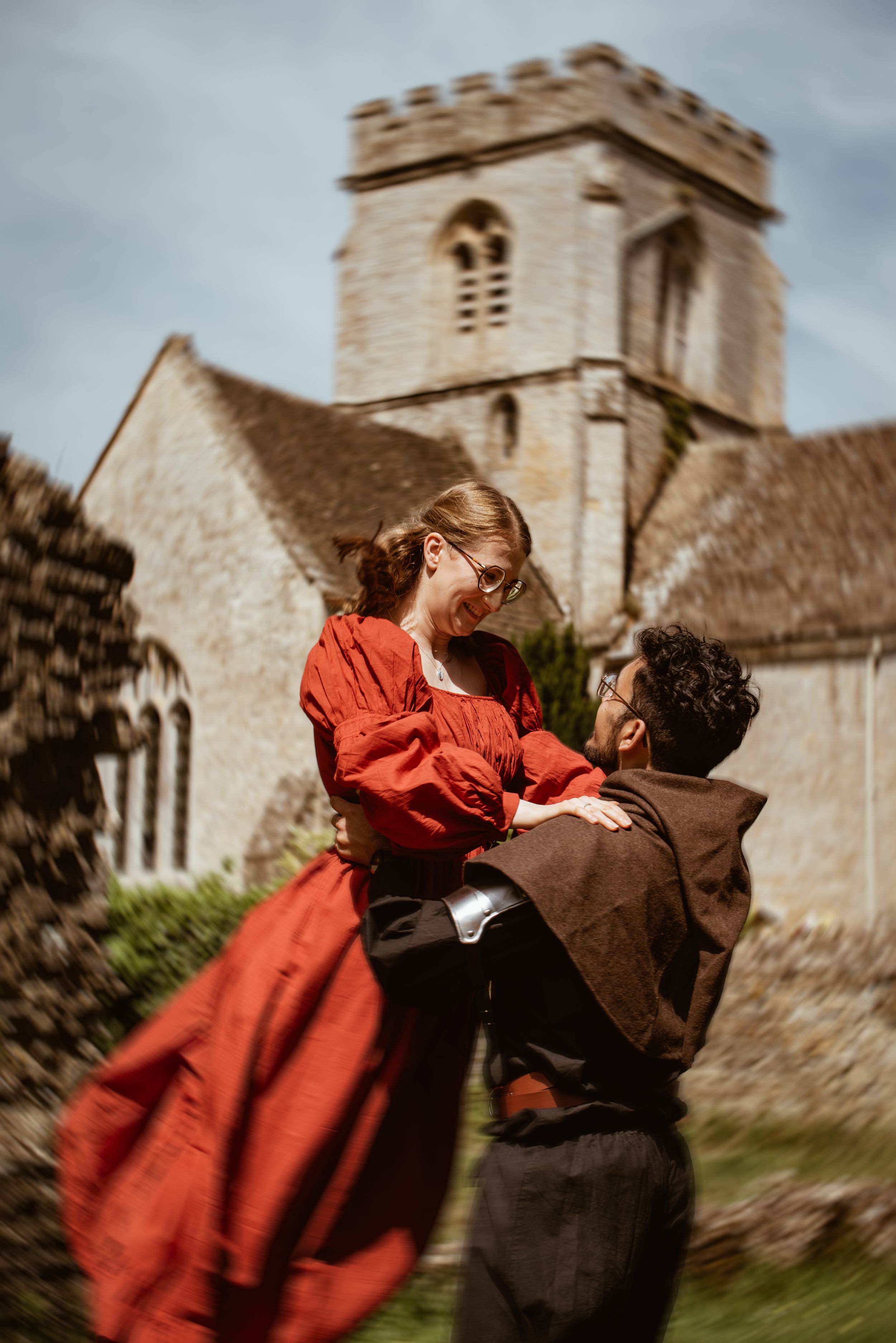 A man in medieval costume lifts a woman in a red dress outside a historic stone church.