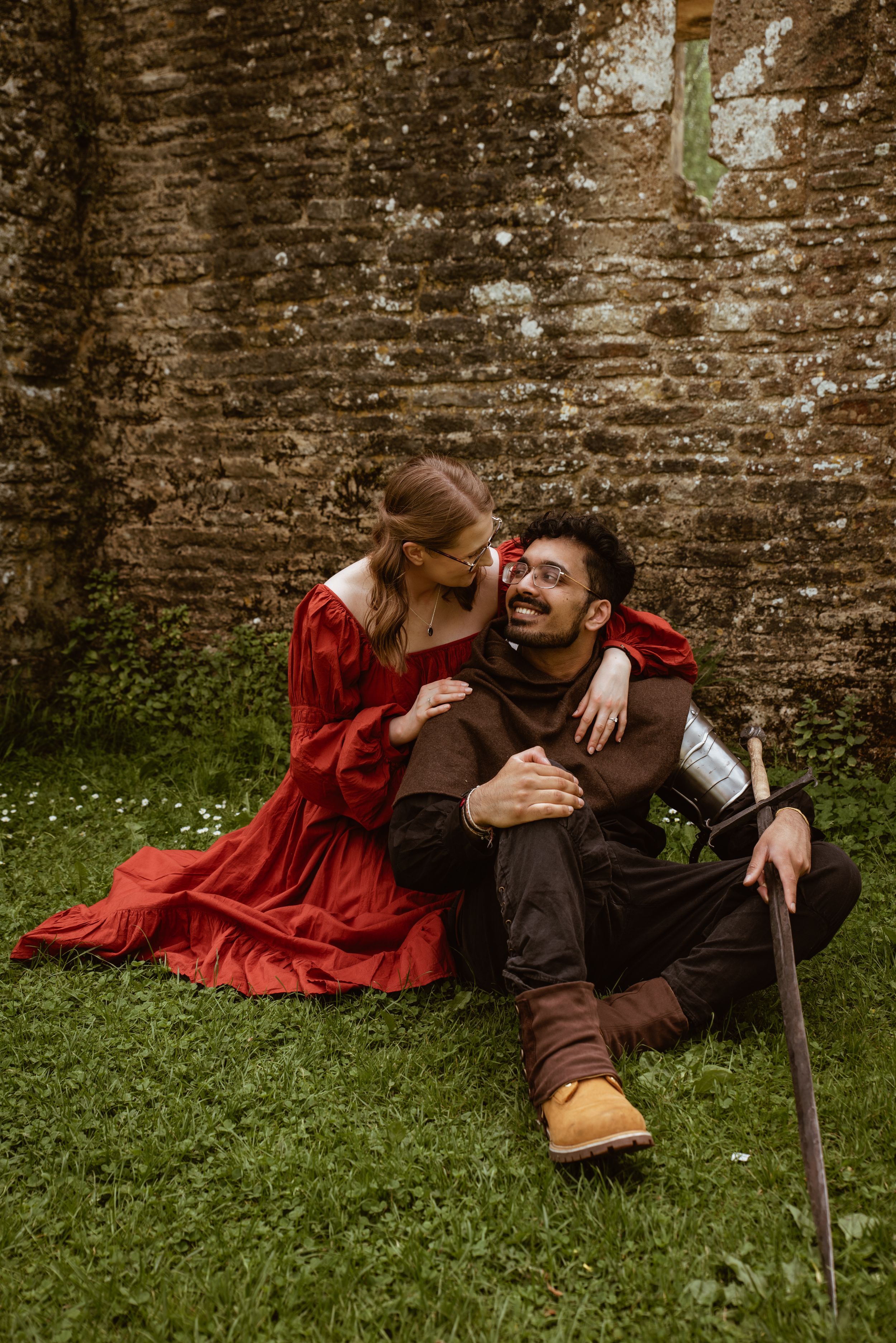 Couple in medieval costumes sitting on grass, woman in red dress whispering to smiling man in brown outfit.
