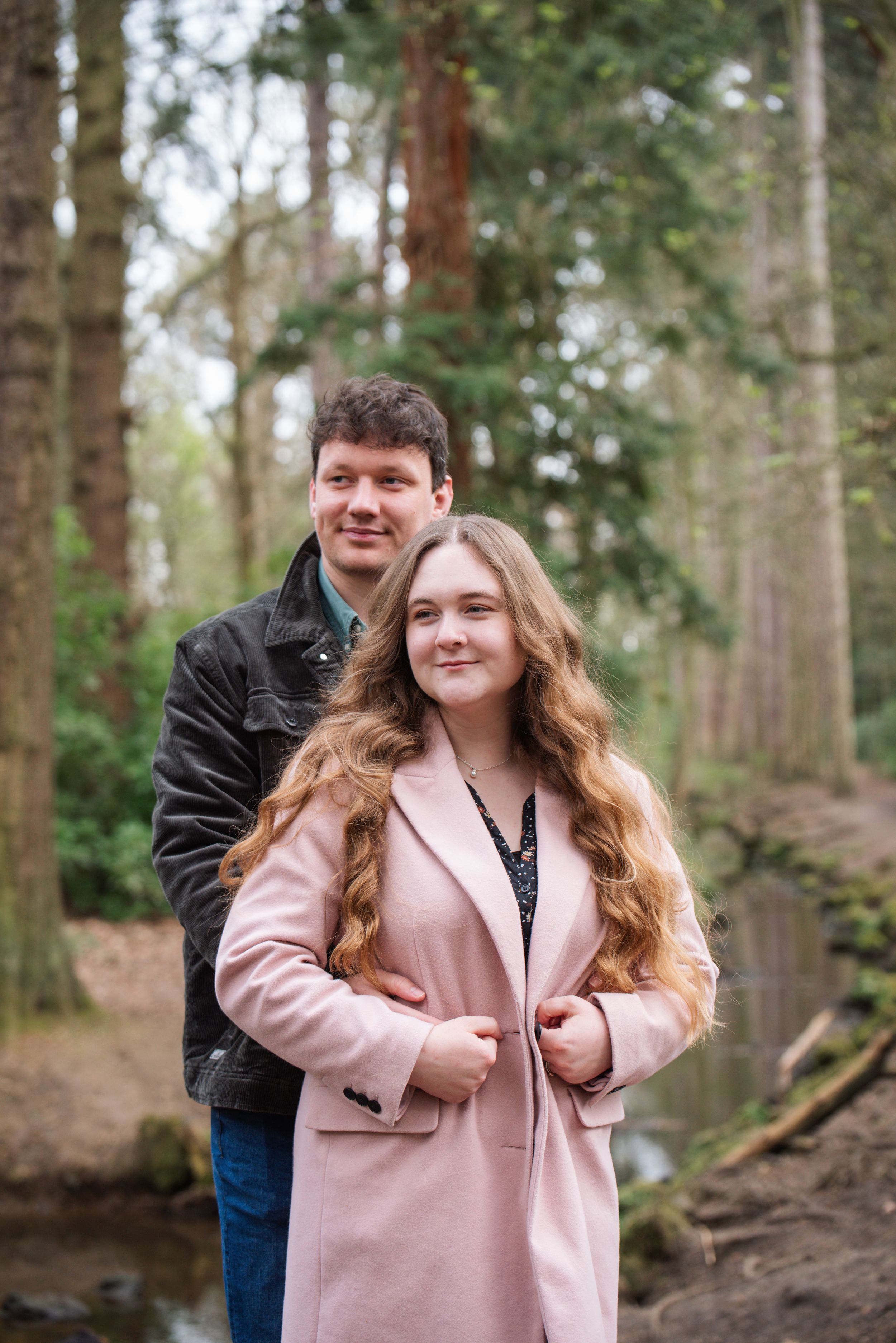 A couple poses together in a forest, woman in pink coat with long wavy hair, man in dark jacket behind her.