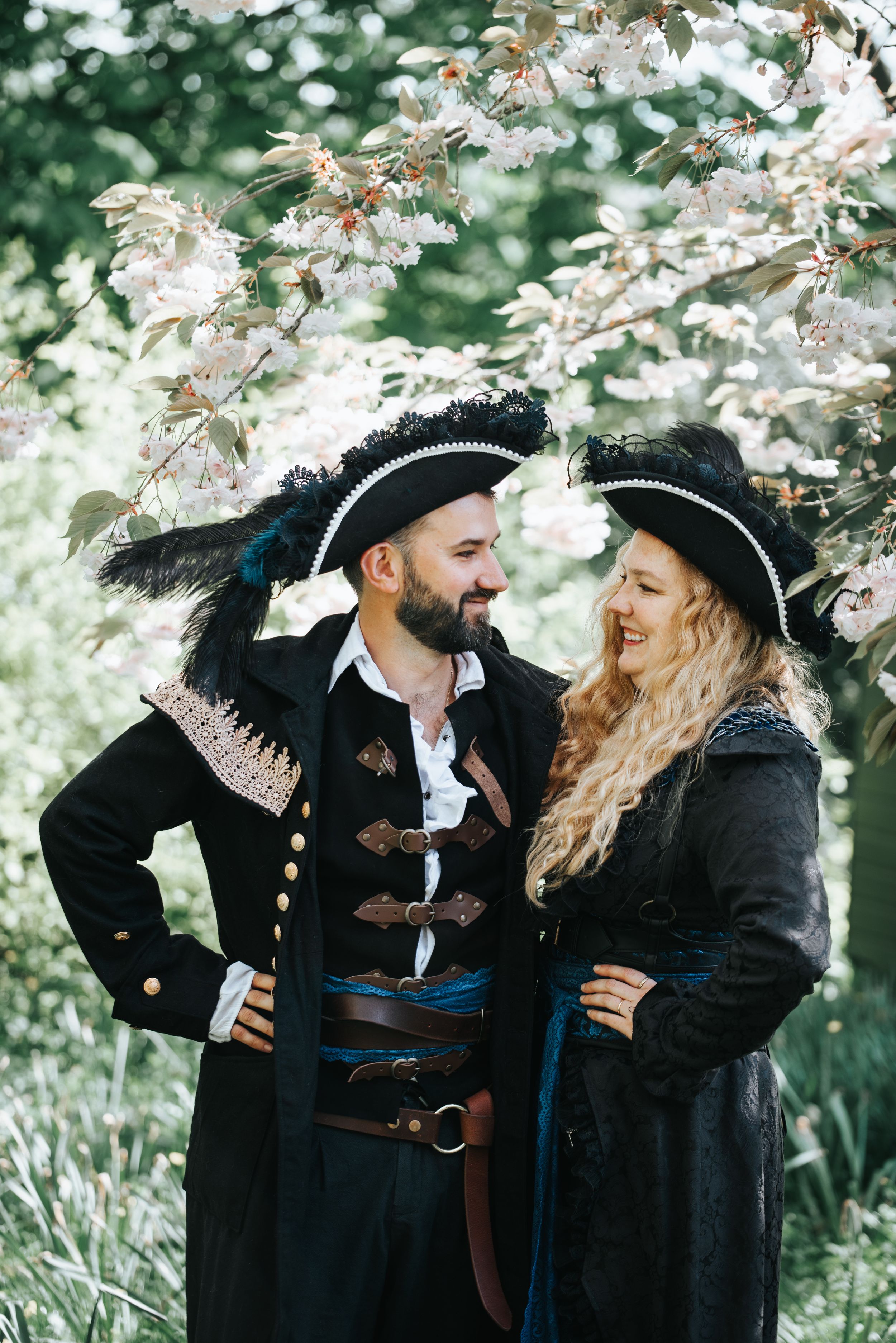 Couple dressed in elegant pirate costumes posing together under flowering trees in a lush garden setting.