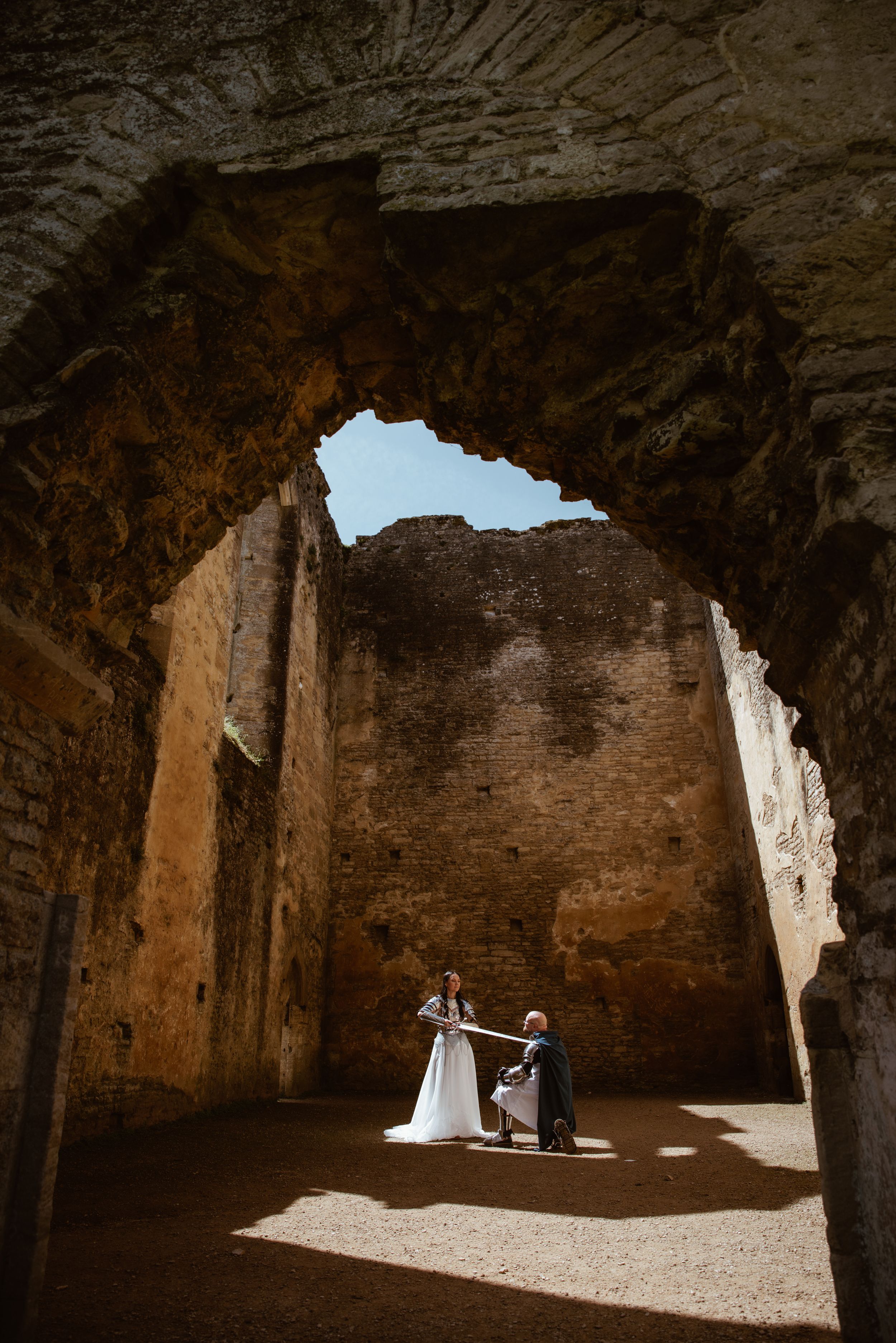 Bride and groom share a romantic moment inside ancient stone ruins, with sunlight streaming through a large arched opening above.