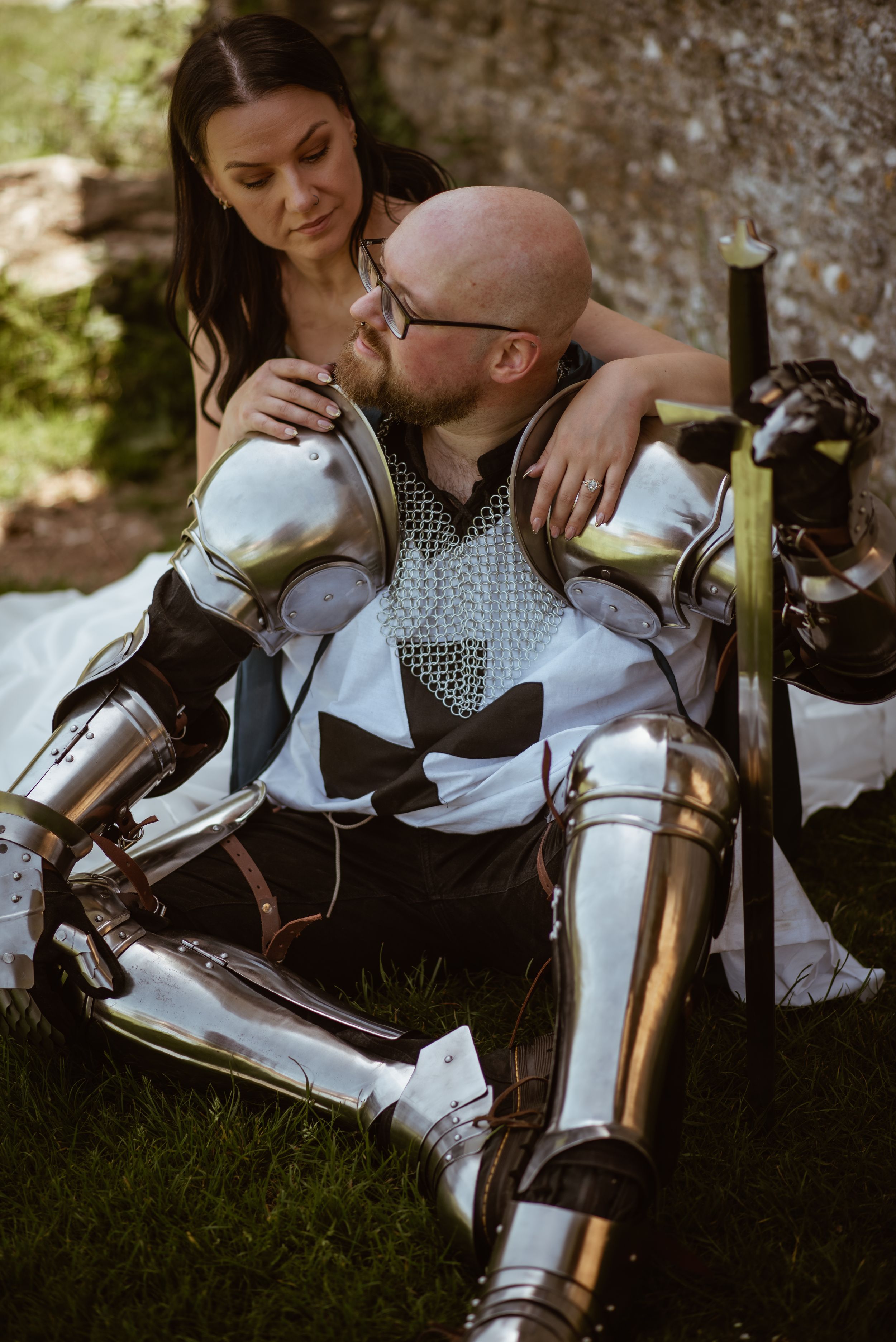 A woman in a dress embraces a bald man wearing medieval knight armor while sitting together on grass outdoors.