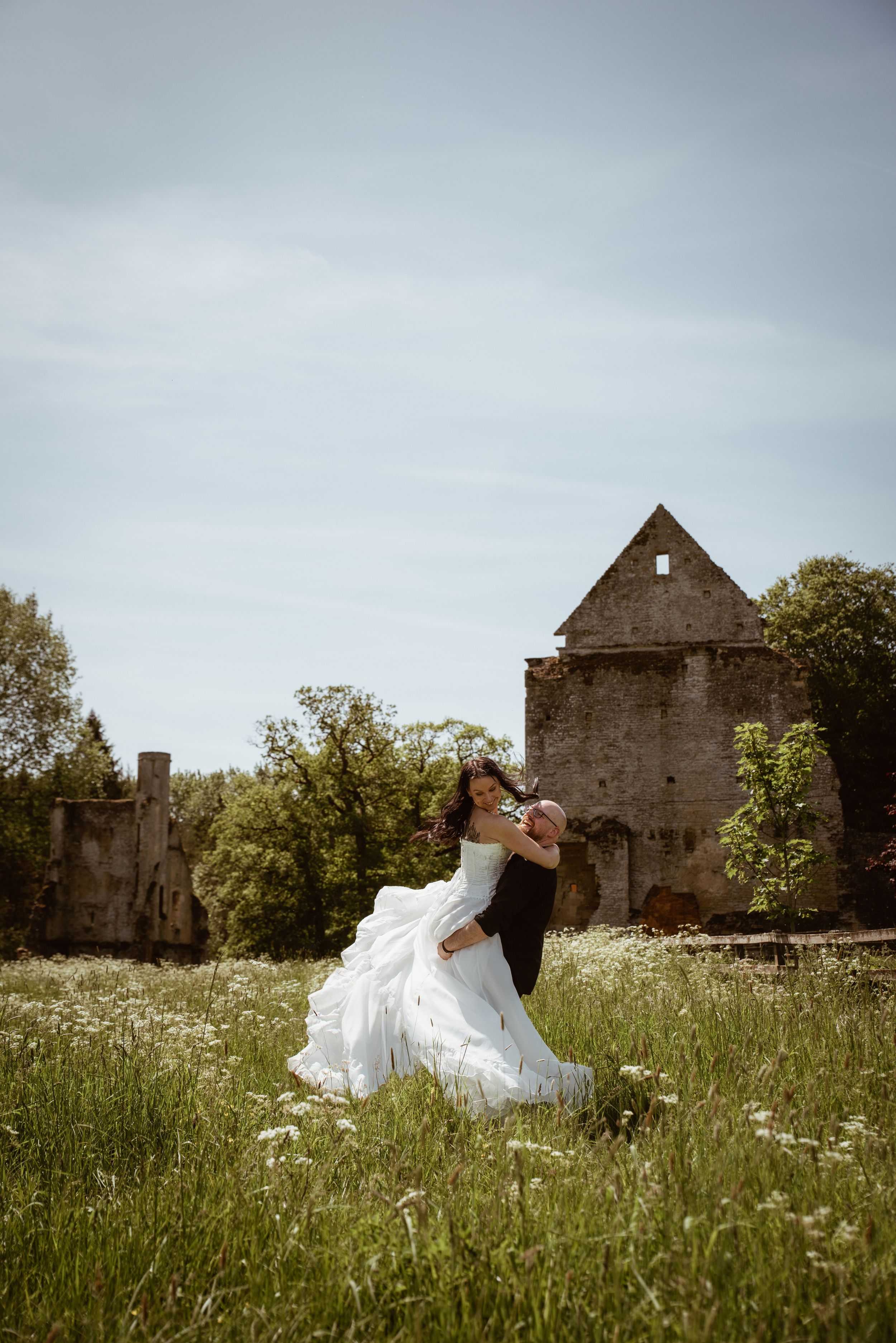 Bride in white gown and groom in black suit dance in a wildflower meadow near rustic stone ruins on a cloudy day.