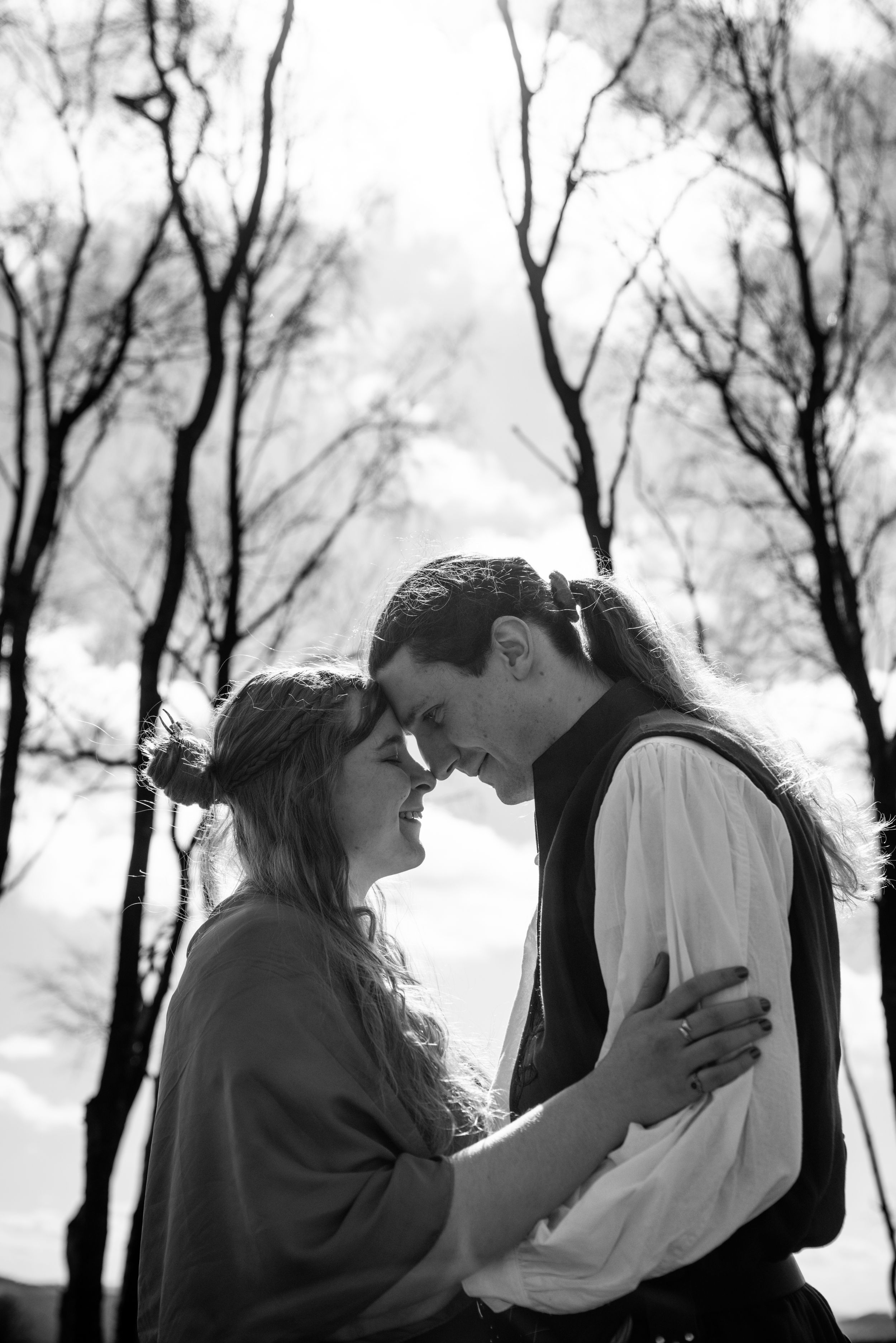 Black and white photo of a romantic couple embracing outdoors with bare trees and cloudy sky in background.