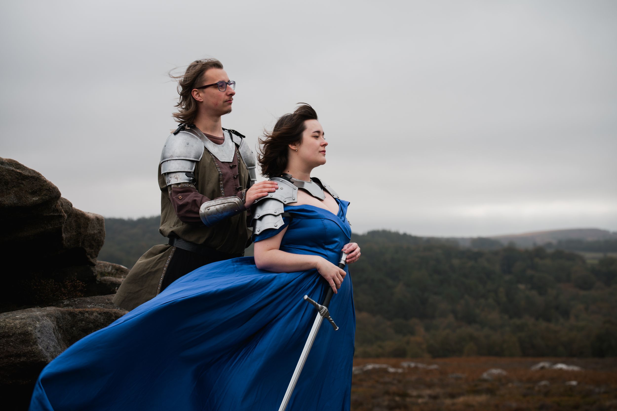 Two people in fantasy outfits pose dramatically against a misty mountain landscape.