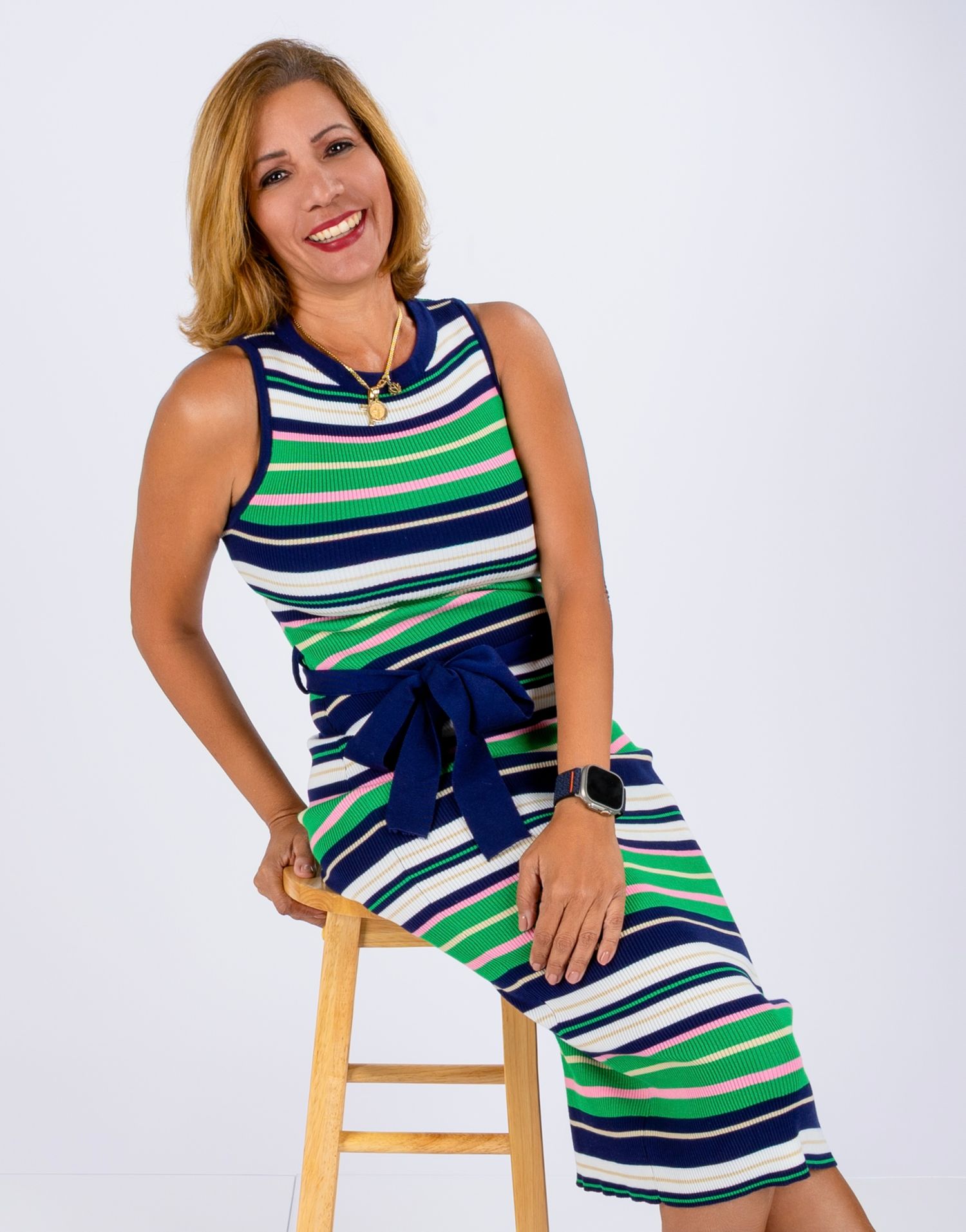 Professional studio portrait of person in striped green and navy dress seated on wooden stool.