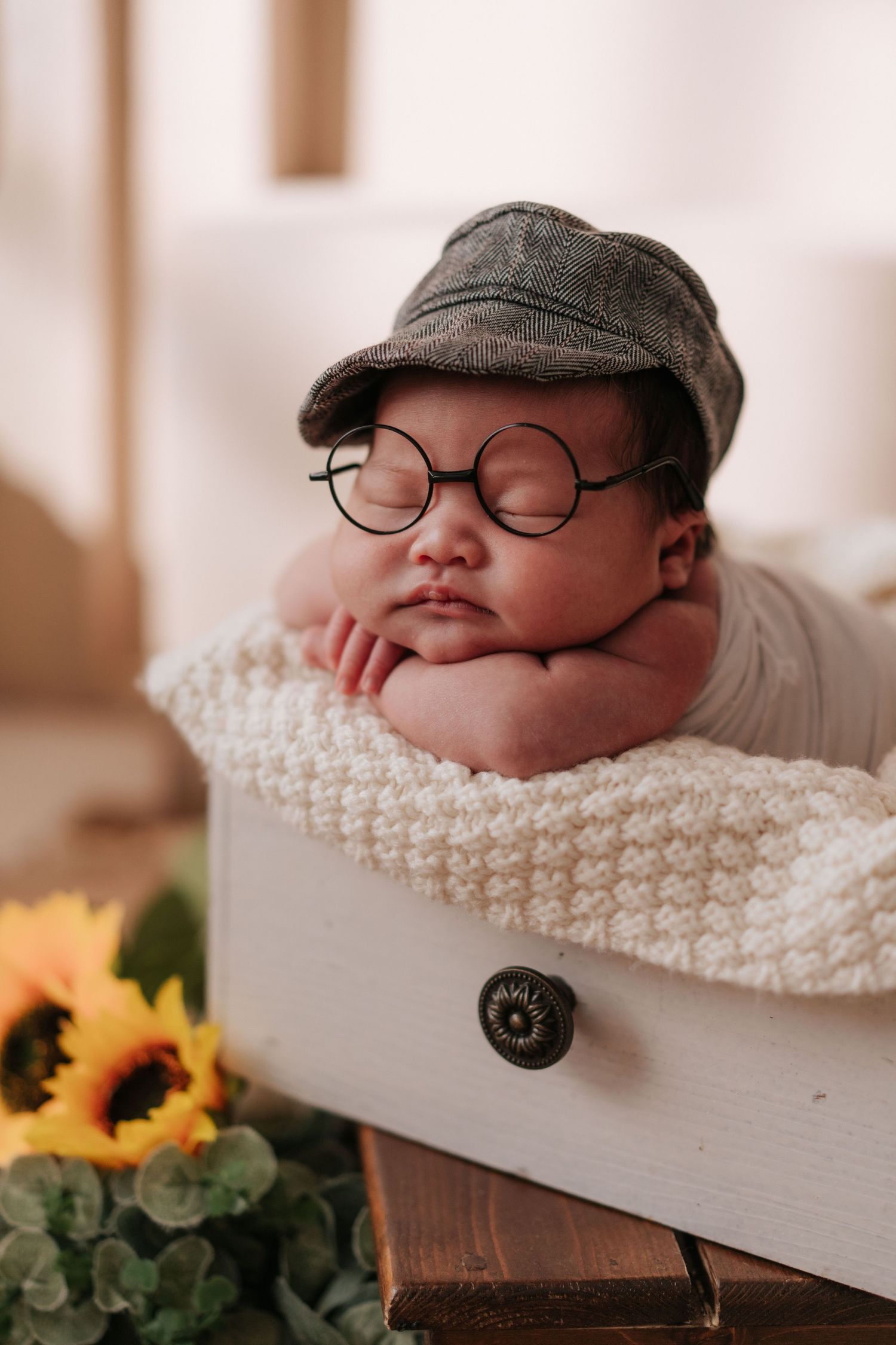 Sleeping newborn wearing vintage style cap and round glasses on knit blanket with sunflowers.