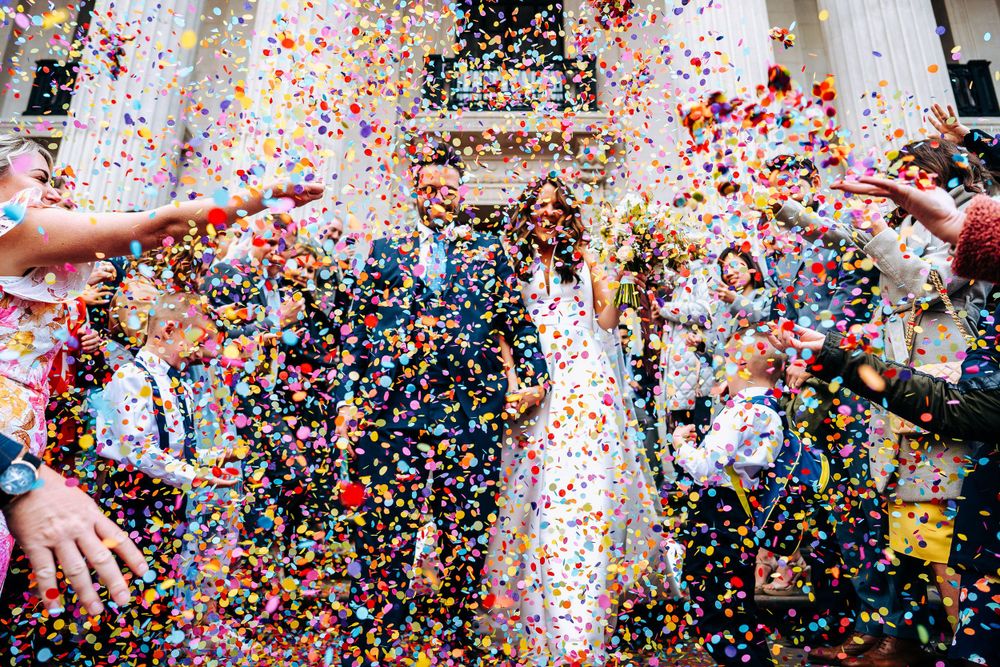 A newlywed couple walks through a shower of colorful confetti thrown by guests outside a grand wedding venue.