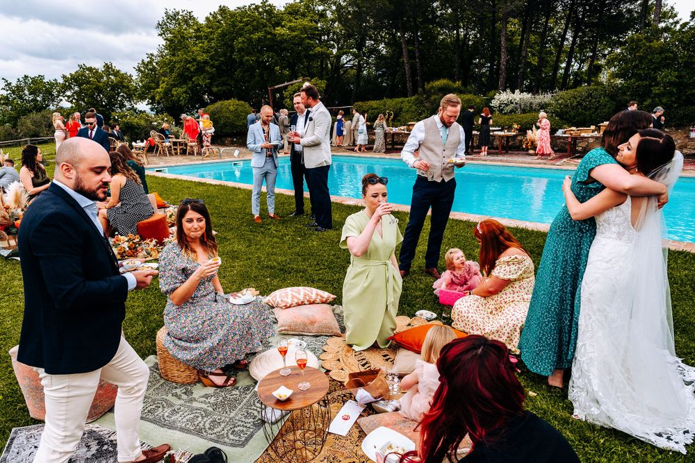 A bride embraces guests during an outdoor wedding reception beside a pool surrounded by colorful decor and greenery.