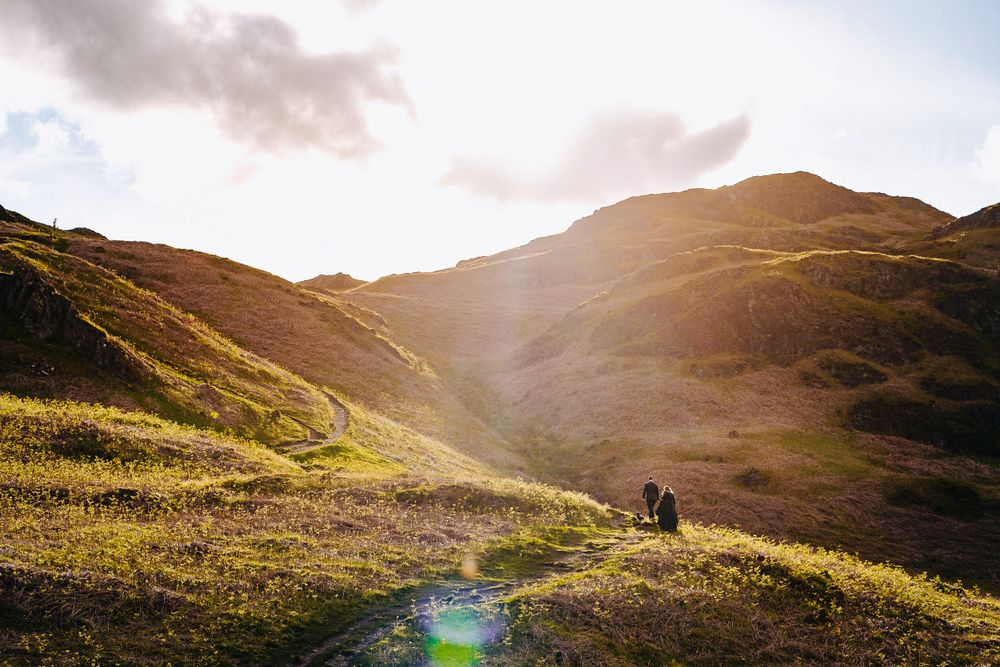 A couple stands together on a sunlit hillside trail surrounded by dramatic rolling green mountains at golden hour.