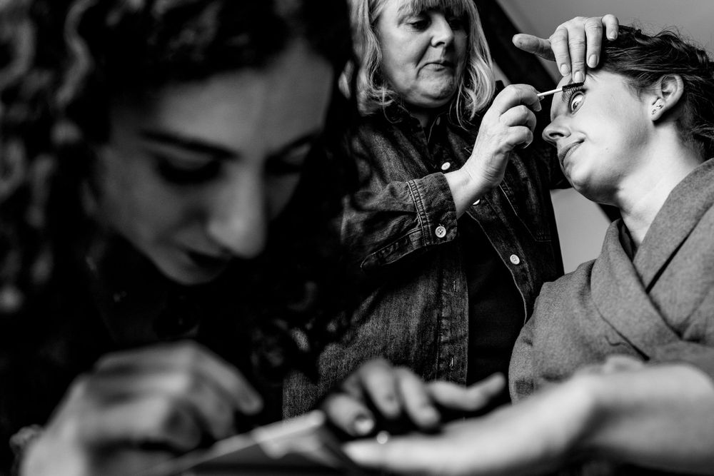 A black and white photo of a makeup artist applying makeup to a bride while another woman reads nearby.