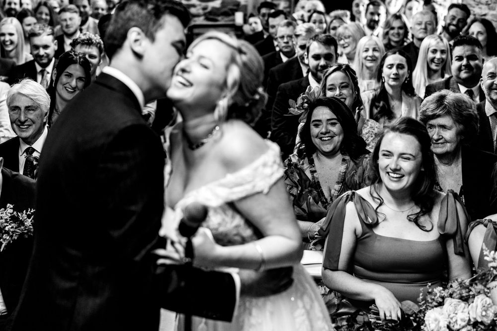 A bride and groom share a joyful moment during their ceremony as smiling wedding guests watch in black and white.