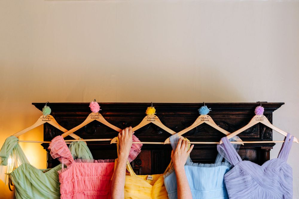 Hands display five colorful bridesmaid dresses on wooden hangers with decorative pineapple clips against a neutral wall.