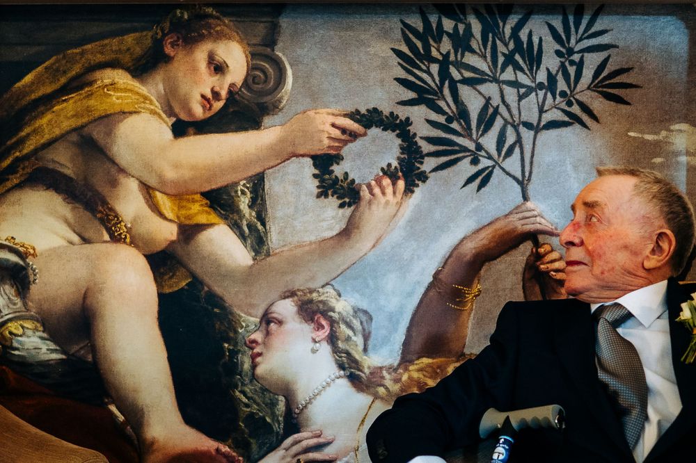 Elderly man posing beside classical painting of women with olive branch in art gallery.