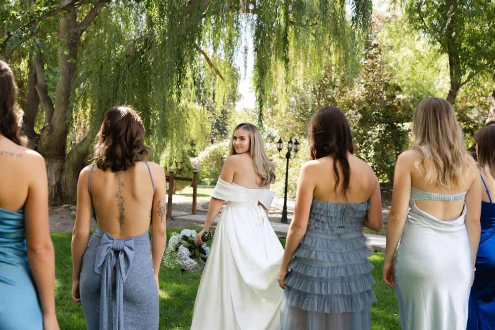 Bridesmaids in blue dresses and bride in white gown standing under willow trees in outdoor garden setting.