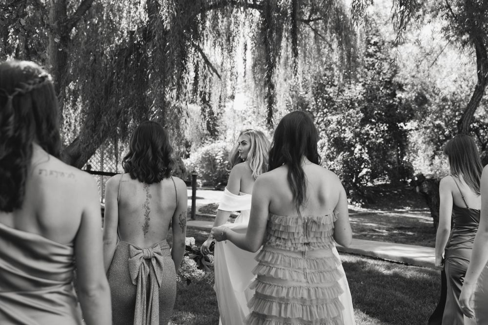 Group of bridesmaids in summer dresses walk through a garden with hanging willow trees in black and white.
