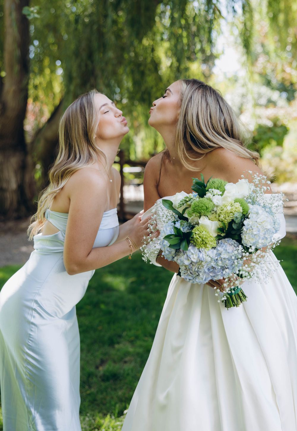 Two people in light dresses share a joyful moment outdoors with a beautiful bouquet of blue and white hydrangeas.