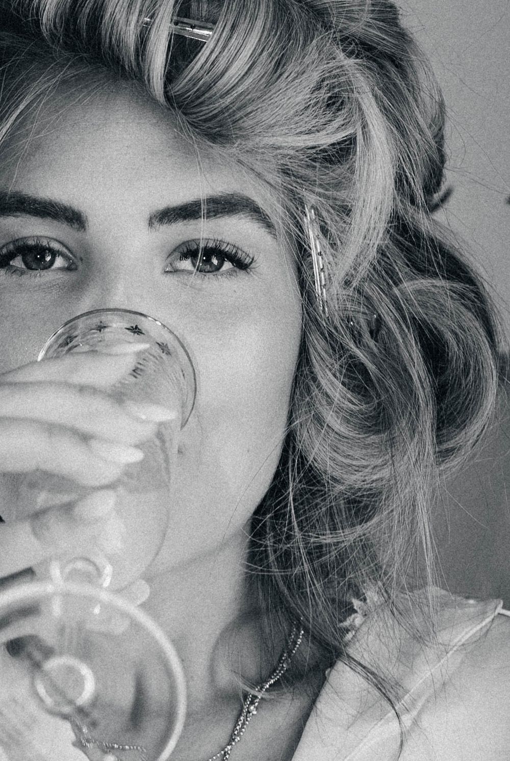 Black and white close-up portrait shows wavy blonde hair and a wine glass held up to partially obscure the view.